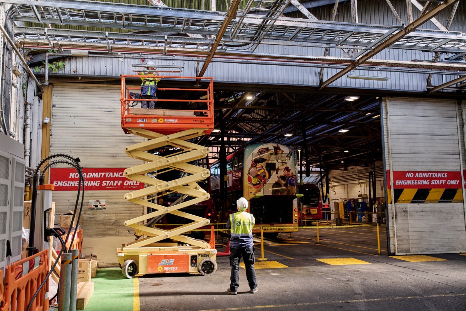 A worker on a scissor lift installs new lighting in a bus garage while another stands below in a hi-vis vest.
