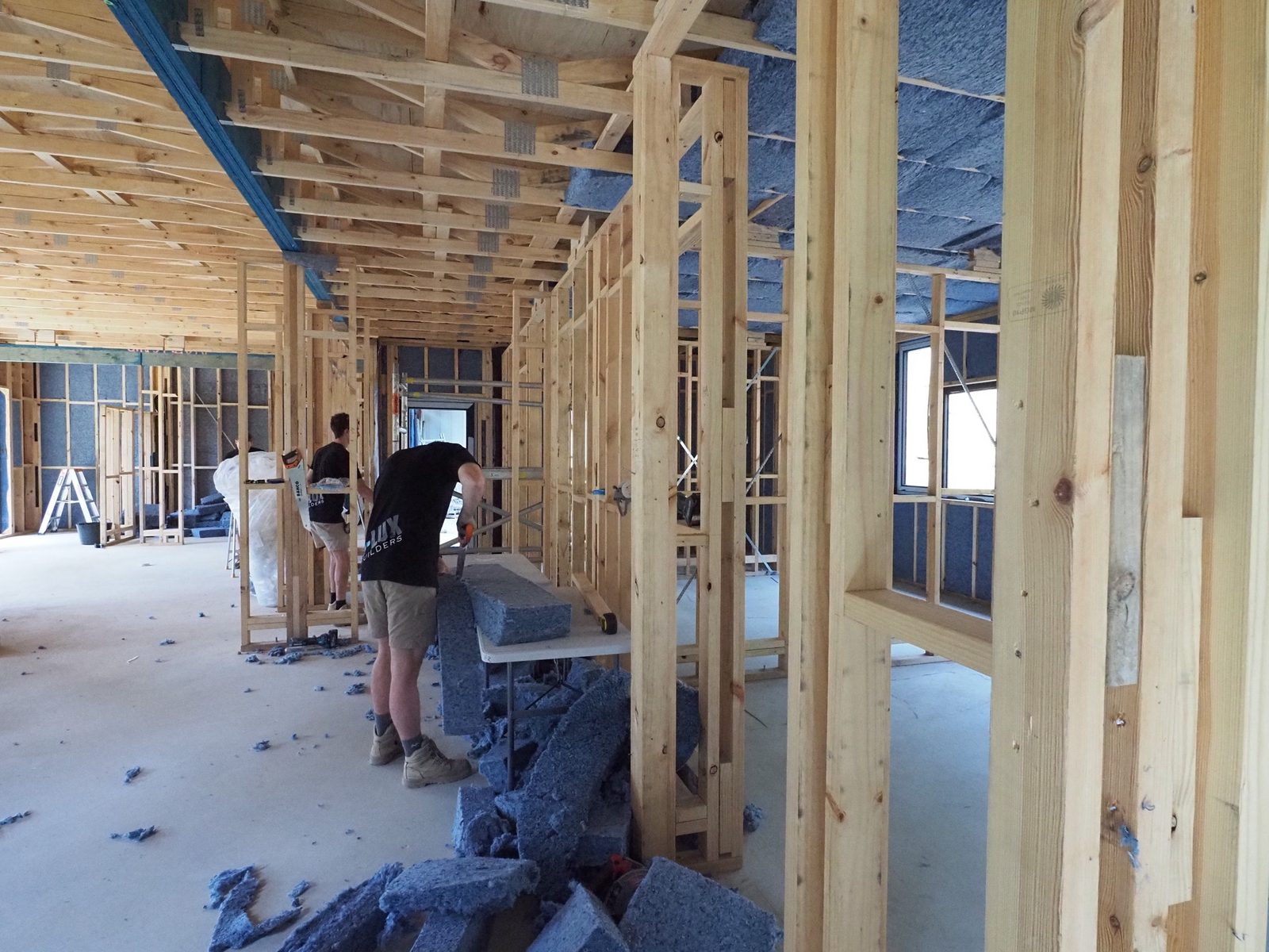 Installers fitting blue-grey recycled denim insulation batts between timber wall studs on a building site.