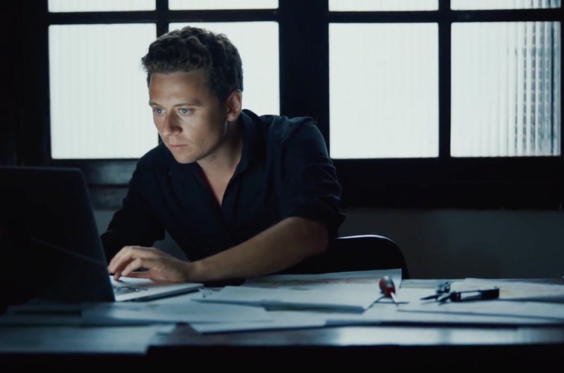 A man in a dark shirt working at a laptop at a desk in front of a large paned window, lit by the screen.