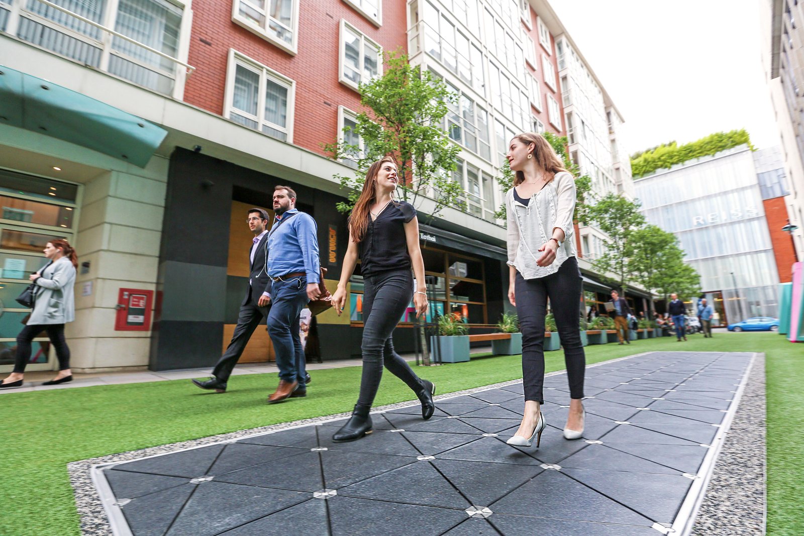 Pedestrians walking along a street beside a strip of triangular Pavegen floor tiles set into the paving, with shops and brick buildings behind.
