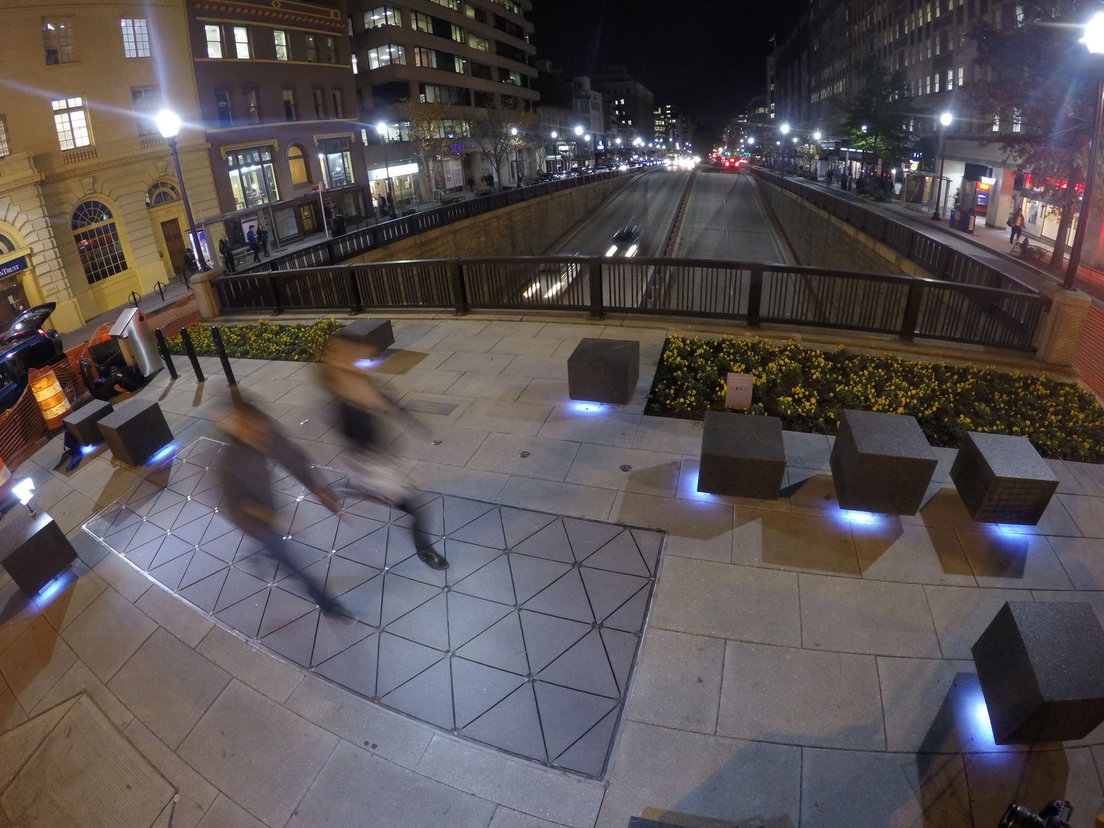 Night view of the pocket park at Dupont Circle in Washington DC, with blurred walkers crossing triangular Pavegen tiles beside lit stone cubes.