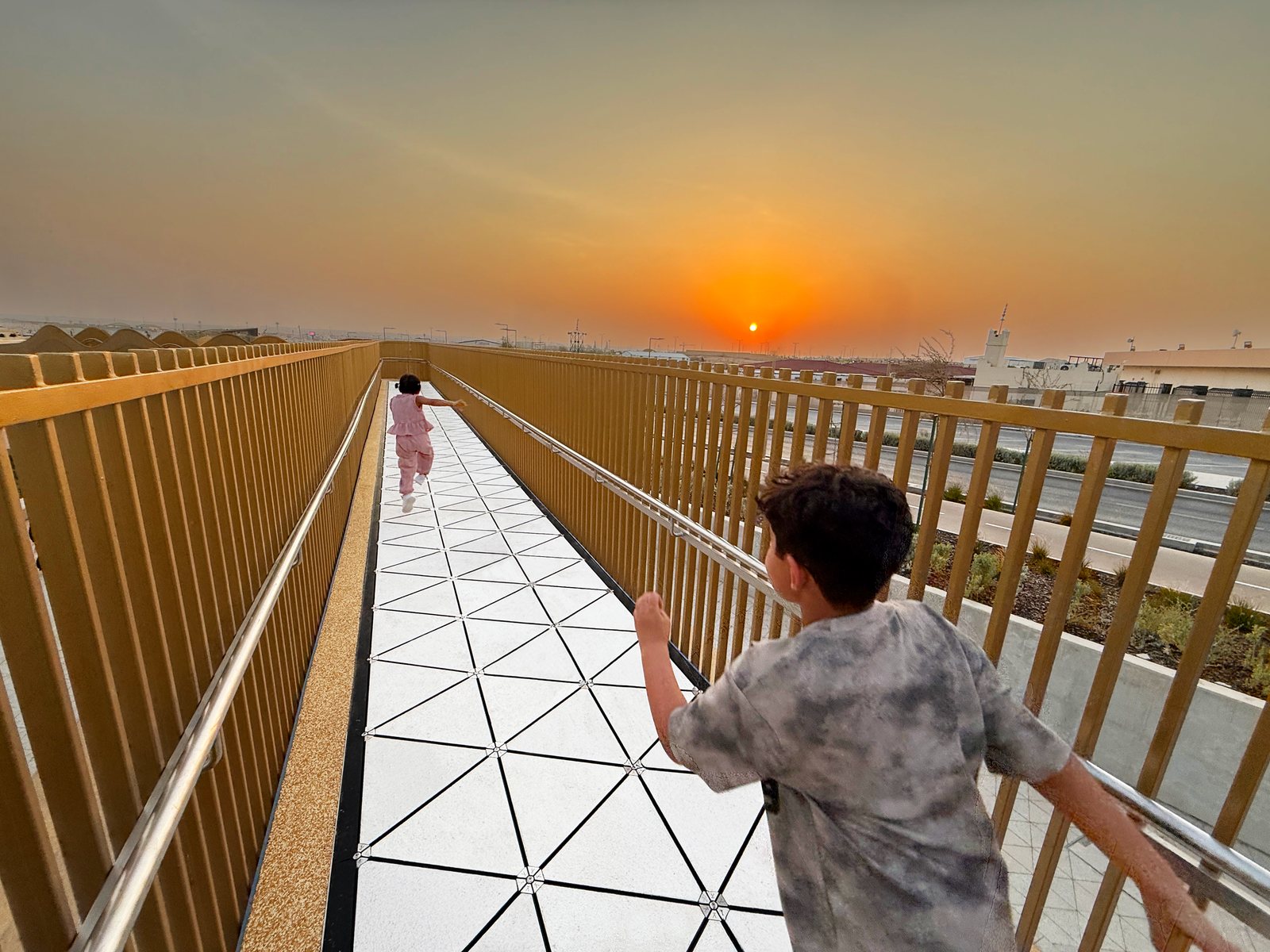 A boy running along a Pavegen walkway of white triangular tiles between golden handrails at sunset in Riyadh.