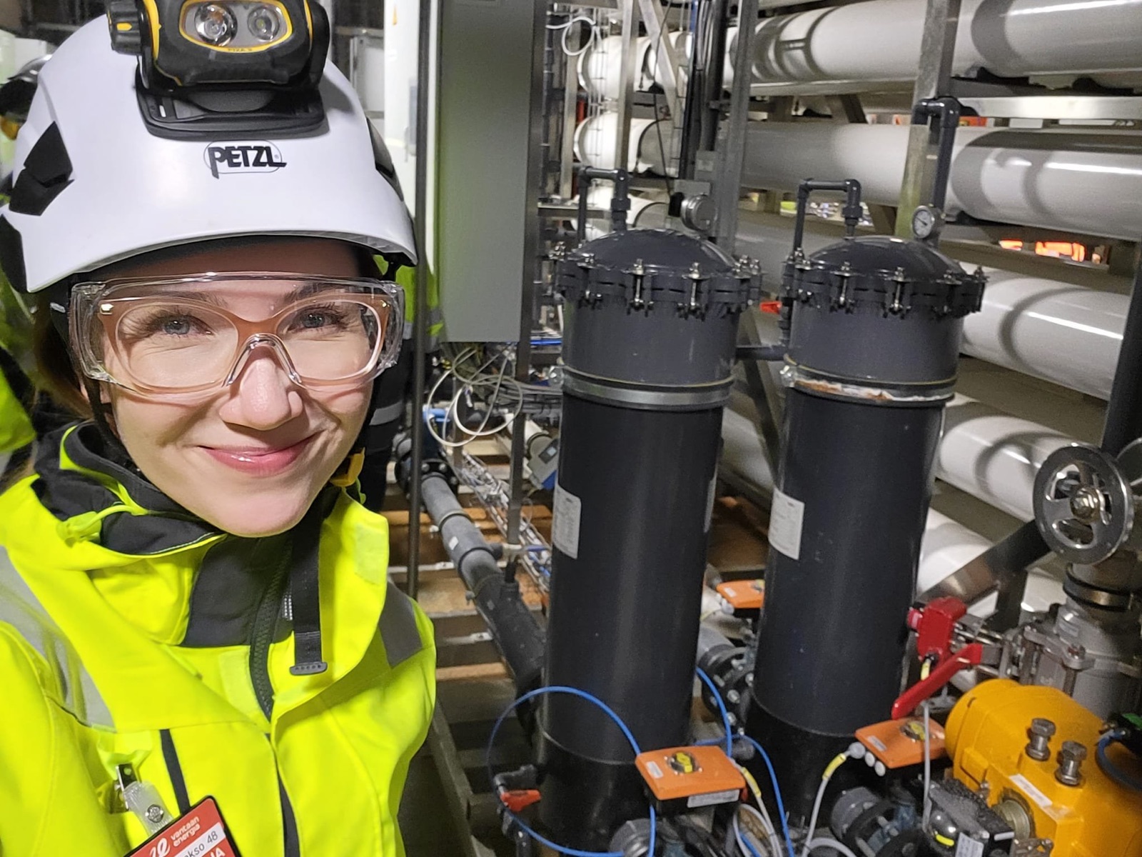 A smiling woman in a white hard hat with a head torch, safety glasses and a yellow high-visibility jacket, standing beside two dark filter vessels inside an industrial plant.