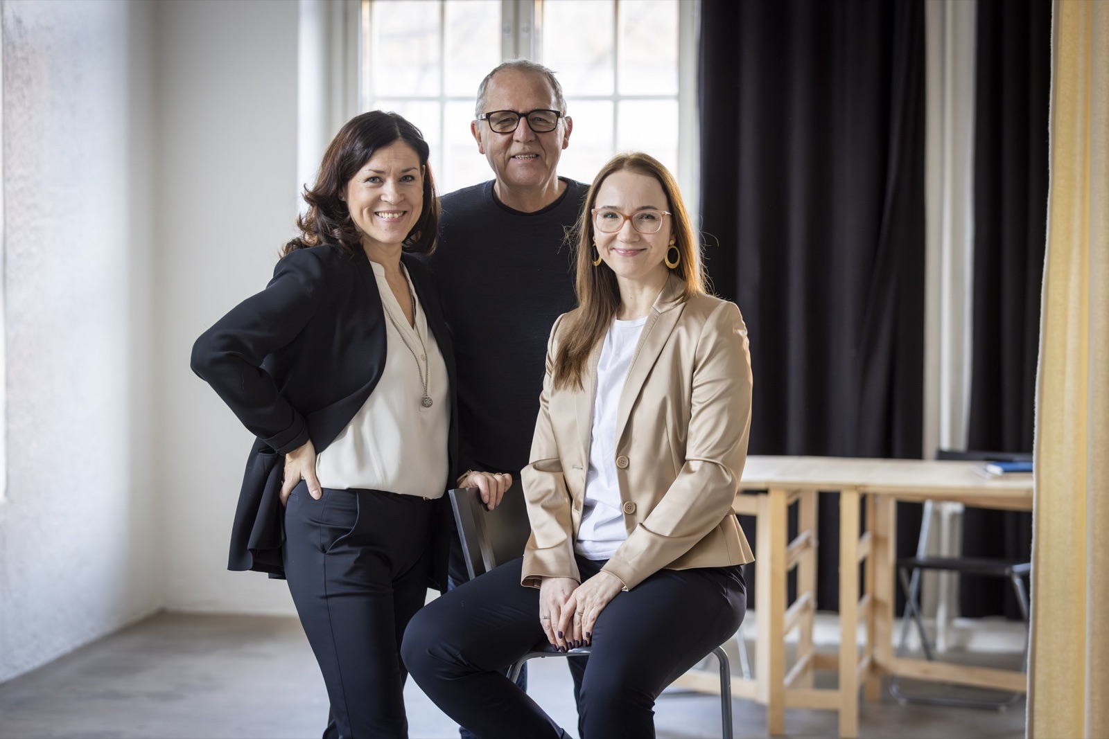 Three people in a bright room with tall windows: a woman in a black blazer, a man in glasses standing behind, and a woman in a beige blazer seated on a stool.