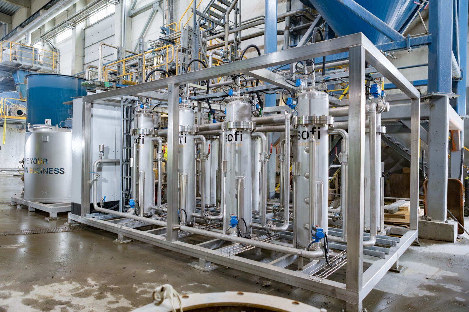 A row of stainless steel Sofi Alchemist filter vessels in a metal frame on the floor of a processing plant, with pipework and a blue tank behind.