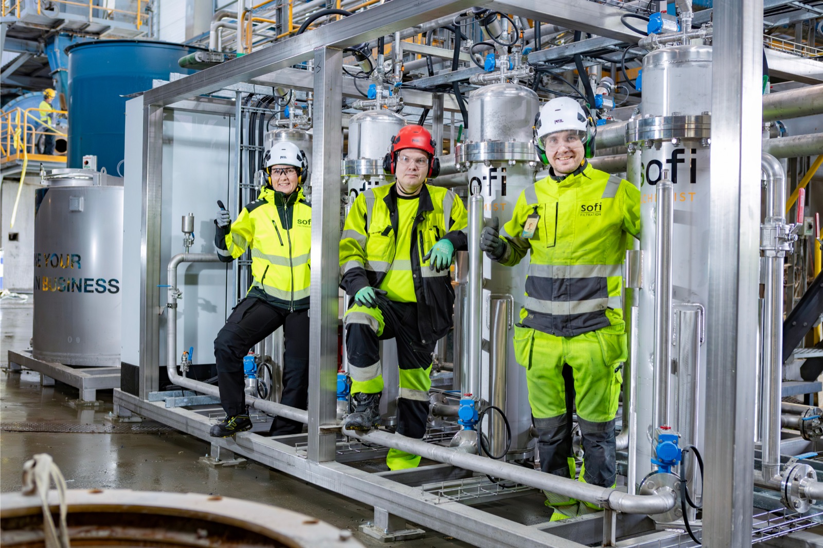Three workers in hard hats and yellow high-visibility gear, two giving a thumbs up, standing inside the frame of the stainless steel Sofi filter unit.