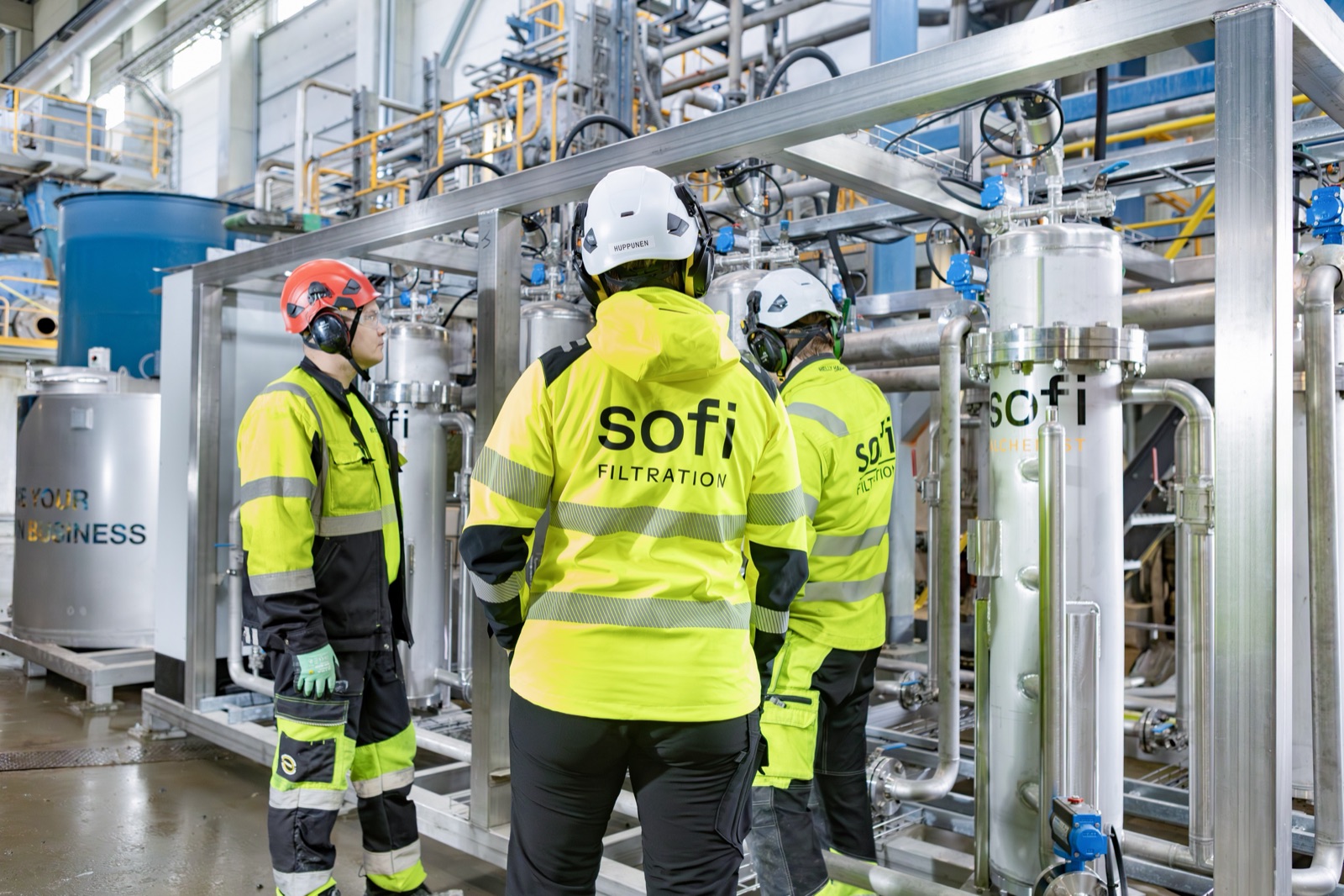Three workers in hard hats and Sofi Filtration jackets, seen from behind, looking at the stainless steel filter vessels of the unit.