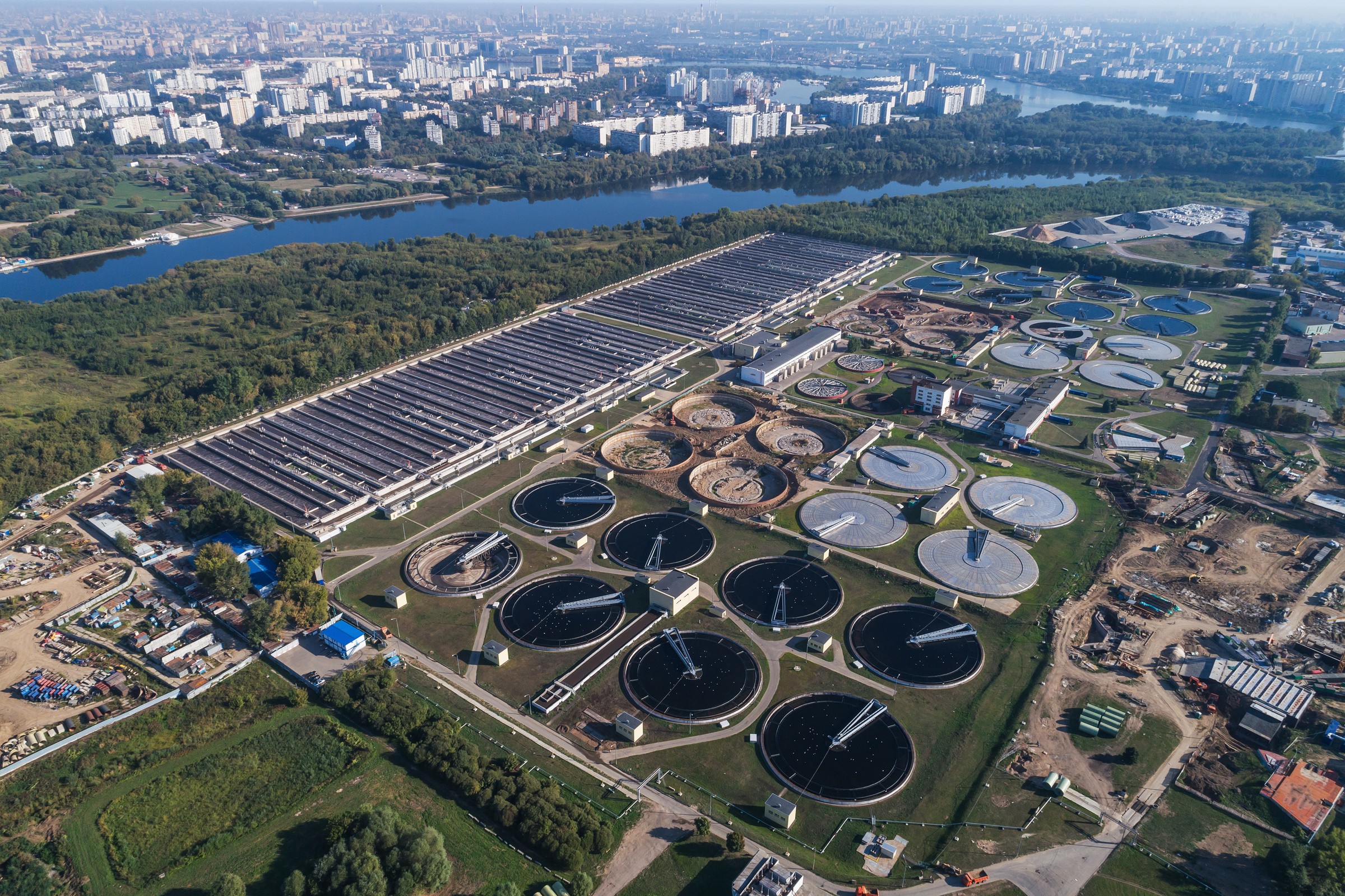 Aerial view of a large wastewater treatment plant beside a river, with round clarifier tanks and long rectangular basins, and a city skyline behind.